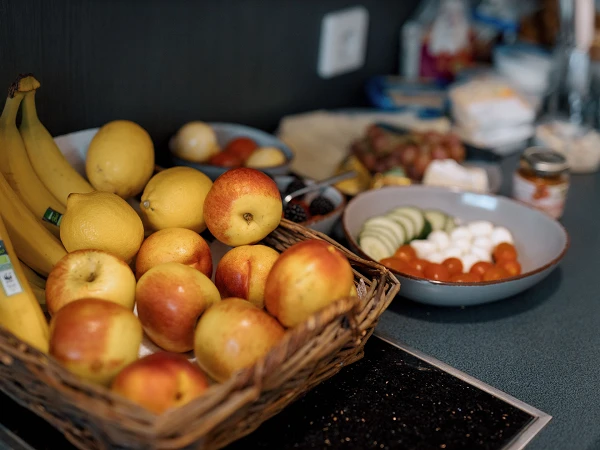 Ein Korb voller Äpfel befindet sich auf einem Küchentisch, daneben liegen Bananen und Zitronen. Im Hintergrund sind verschiedene weitere Lebensmittel wie Trauben, Gurkenscheiben, Kirschtomaten und Käsebällchen auf Tellern und in Schalen angeordnet.