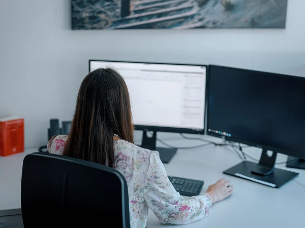 Eine Person mit langen, glatten Haaren sitzt mit dem Rücken zur Kamera an einem Schreibtisch und arbeitet an einem Computer mit zwei Monitoren. Auf dem Schreibtisch sind einige Büromaterialien und ein rotes Objekt sichtbar.