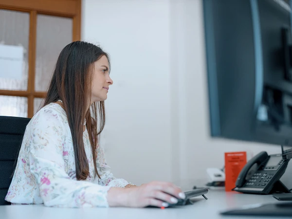 Eine Frau mit langen, braunen Haaren sitzt an einem Schreibtisch und konzentriert sich auf einen Computerbildschirm, während sie die Maus in der Hand hält. Am Schreibtisch befinden sich ein Telefon und einige Dokumente.
