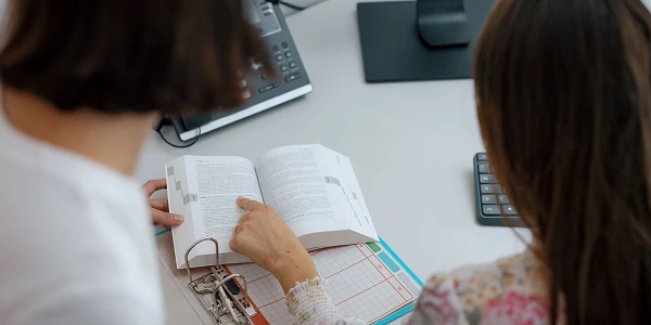 Zwei Personen sitzen an einem Schreibtisch und betrachten gemeinsam ein aufgeschlagenes Buch, während eine von ihnen mit dem Finger auf eine Textstelle zeigt. Im Hintergrund ist ein Computer mit einem geöffneten Dokument zu sehen.
