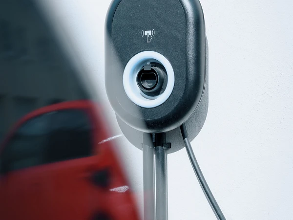 Eine elektrische Ladestation für Fahrzeuge, montiert an einer Wand, mit einem Ladekabel, das bereit ist, in ein Elektroauto eingesteckt zu werden. Im Hintergrund ist teilweise ein rotes Auto sichtbar.