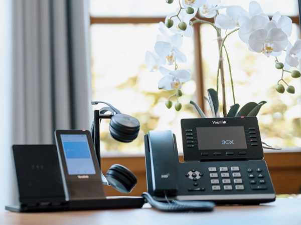 Ein moderner Schreibtisch mit einem VoIP-Telefon, einem Headset und einem kleinen Bildschirm auf der linken Seite. Im Hintergrund sind Fenster mit hellen Vorhängen und ein Topf mit weißen Orchideen zu sehen.