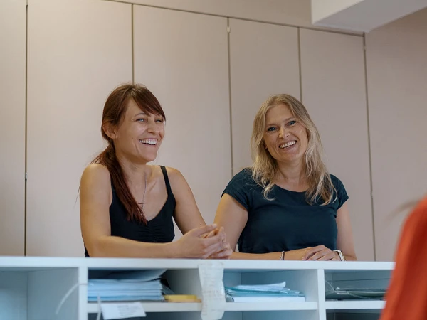 Zwei Frauen stehen an einem Empfangstresen und lachen, während sie mit einer anderen Person sprechen, die auf der gegenüberliegenden Seite sitzt. Der Schreibtisch ist mit verschiedenen Papierunterlagen und Büromaterialien bedeckt.