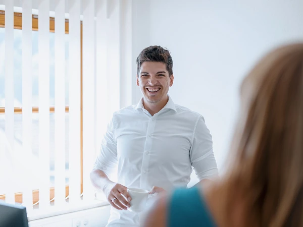Ein Mann mit dunklen Haaren trägt ein weißes Hemd und hält eine Tasse in der Hand, während er lächelt und in Richtung einer anderen Person blickt. Im Hintergrund sind Jalousien und ein Fenster zu sehen.