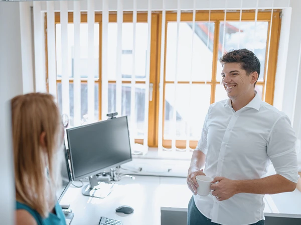 Ein Mann in einem weißen Hemd lächelt, während er in einem hellen Büro mit einem Becher in der Hand mit einer Frau spricht, die ihm gegenübersteht. Im Hintergrund sind ein Computer und Fenster mit Jalousien zu sehen.