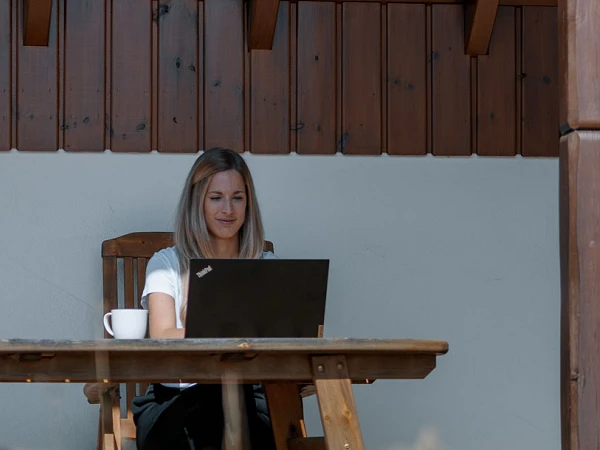 Eine Frau sitzt entspannt in einer schaukelnden Liege unter einem Dach und arbeitet auf einem Laptop, während eine andere Frau am Tisch daneben mit einem Laptop arbeitet und eine Tasse Kaffee vor sich hat.