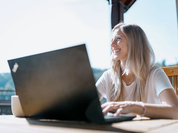 Eine lachende Frau mit langen, blonden Haaren sitzt an einem Tisch im Freien und arbeitet mit einem Laptop, während sie die Aussicht auf eine bewaldete Landschaft genießt.