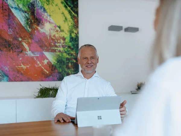 Ein Mann in einem weißen Hemd sitzt an einem Tisch und lächelt, während er ein Tablet in den Händen hält. Im Hintergrund ist ein buntes, abstraktes Kunstwerk und eine Tasse Kaffee auf dem Tisch sichtbar.