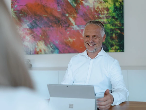 Ein lächelnder Mann in einem weißen Hemd sitzt an einem Tisch und benutzt ein Tablet, während er einem Gesprächspartner gegenüber sitzt. Im Hintergrund hängt ein buntes abstraktes Kunstwerk.