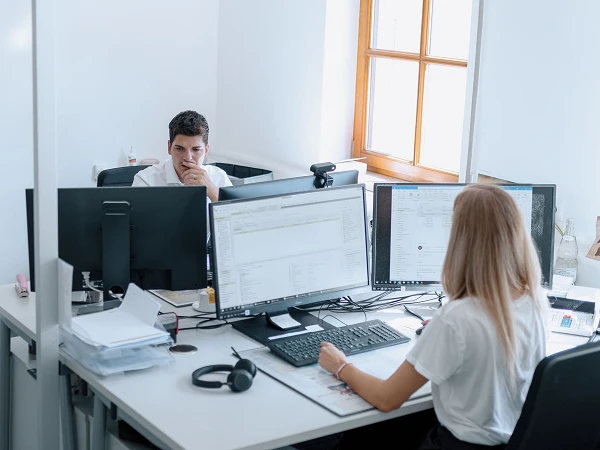 Ein modernes Büro mit zwei Mitarbeitern; einer sitzt am Schreibtisch mit mehreren Monitoren und arbeitet, während der andere am Fenster steht und nachdenklich auf seinen Bildschirm schaut.
