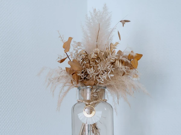 Ein Glasvase steht auf einem weißen Tisch und enthält getrocknete Blumen und Pflanzen in sanften Erdtönen. Die Wand im Hintergrund ist in einem schlichten, strukturierten Weiß gehalten.