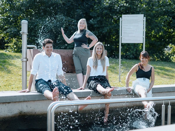 Vier Personen sitzen auf dem Rand eines Wasserbeckens und halten die Füße im Wasser, während eine der Frauen im Hintergrund freundlich in die Kamera lächelt und die Hände hebt. Es ist ein sonniger Tag, umgeben von grüner Landschaft.