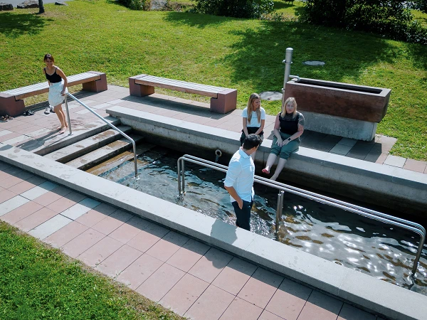 Vier Personen sind in einer Parkanlage zu sehen, wo sie in einem flachen Wasserlauf sitzen oder stehen. Zwei Frauen sitzen am Rand des Wassers, während eine Frau auf einer Treppe zum Wasser hinuntergeht und ein Mann im Wasser steht. Die Umgebung ist grün und gepflegt, mit Bänken aus Holz und Beton.