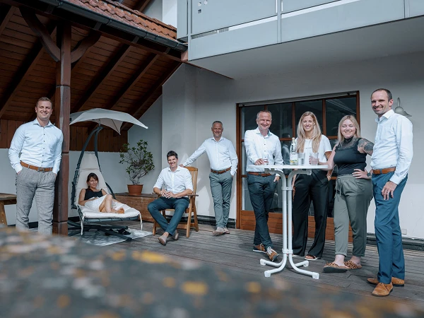 Eine Gruppe von sieben Personen steht und sitzt auf einer Terrasse vor einem Wohnhaus, einige in entspannter Pose, während andere in ein Gespräch vertieft sind. Es herrscht eine freundliche und gesellige Atmosphäre im Freien.