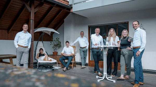 Eine Gruppe von sieben Personen steht und sitzt auf einer Terrasse vor einem Wohnhaus, einige in entspannter Pose, während andere in ein Gespräch vertieft sind. Es herrscht eine freundliche und gesellige Atmosphäre im Freien.