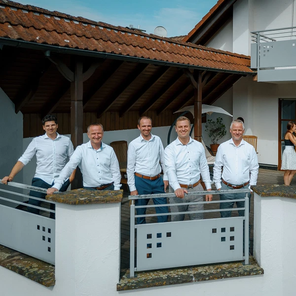 Fünf Männer in weißen Hemden stehen auf einem Balkon mit Blick auf eine Terrasse, auf der mehrere Frauen in lässiger Kleidung Gespräche führen. Im Hintergrund ist ein Gebäude mit einem Dach und einer modernen Außenarchitektur sichtbar.