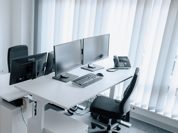 Ein moderner Büroarbeitsplatz mit einem höhenverstellbaren Schreibtisch, zwei Computerbildschirmen, einer Tastatur, einer Maus und einem Telefon, umgeben von hellen Wänden und einem Teppichboden. Die großen Fenster im Hintergrund sind mit weißen Jalousien versehen, die das Licht filtern.