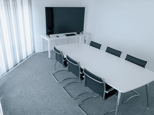 Ein leeres Besprechungszimmer mit einem länglichen weißen Tisch und mehreren schwarzen Stühlen. An der Wand befindet sich ein großer Bildschirm, neben dem ein Tisch mit weiteren Geräten steht. Große Fenster mit vertikalen Jalousien lassen Tageslicht herein.