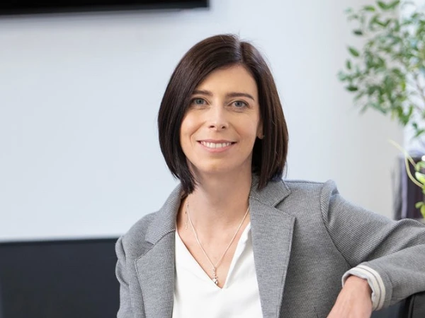 Eine Frau mit schulterlangem, braunem Haar sitzt entspannt in einem modernen Büro, trägt einen grauen Blazer über einem weißen Oberteil und lächelt freundlich in die Kamera. Im Hintergrund ist ein Fernseher und eine Pflanze sichtbar.
