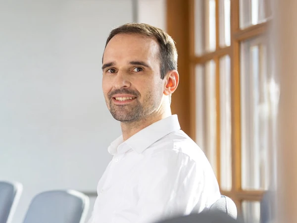 Ein Mann mit kurzen, braunen Haaren und einem freundlichen Lächeln sitzt in einem hellen, modernen Büro. Im Hintergrund befindet sich eine grüne Pflanze in einem Blumentopf und einige Stühle.