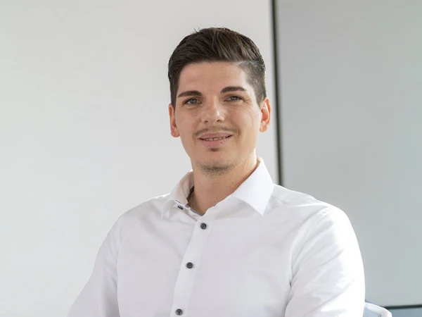 Ein junger Mann mit dunklen Haaren sitzt entspannt in einem modernen Büro, trägt ein weißes Hemd und lächelt leicht in die Kamera. Der Hintergrund ist hell und minimalistisch gestaltet.