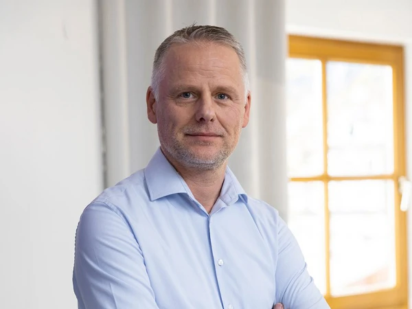 Ein Mann mit kurzen, grauen Haaren steht mit verschränkten Armen vor einem großen Fenster in einem modernen Büro, das mit neutralen Farben und Pflanzen dekoriert ist.