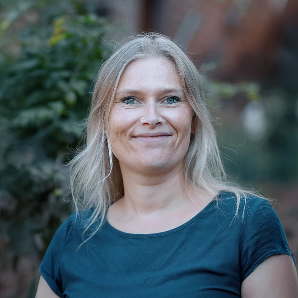 Eine lächelnde Frau mit langen, blonden Haaren steht vor einem unscharfen, grünen Hintergrund. Sie trägt ein schlichtes, dunkelblaues Oberteil und schaut direkt in die Kamera.