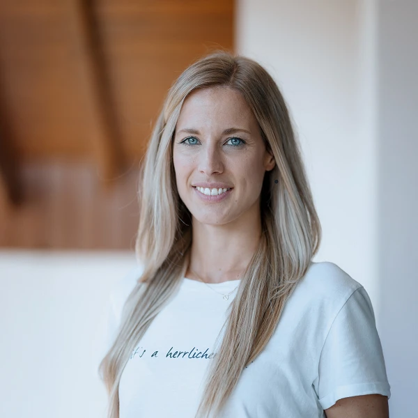 Eine blonde Frau mit langen, glatten Haaren lächelt in die Kamera, während sie ein schlichtes, weißes T-Shirt trägt. Der Hintergrund zeigt einen hellen, minimalistischen Raum mit Holzstrukturen.