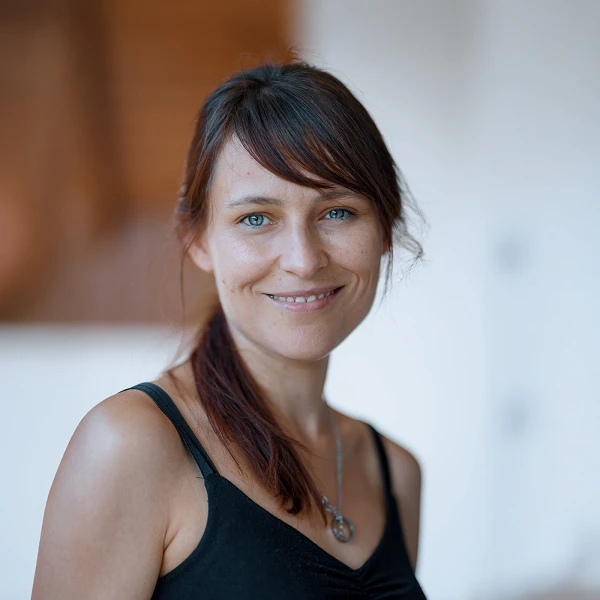 Eine Frau mit langen, braunen Haaren und blauen Augen lächelt freundlich in die Kamera. Sie trägt ein schwarzes Top und steht in einem hellen Raum mit unscharfem Hintergrund.